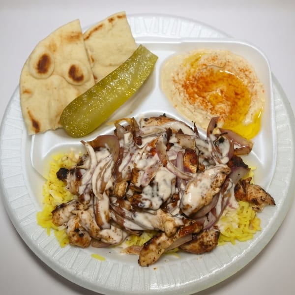 Chicken shawarma plate with rice, hummus, pita bread, and a pickle spear on a white plate.